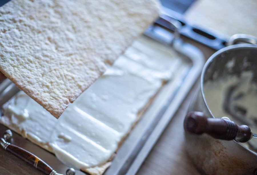 A layer of cake is being placed onto a cream-covered base layer in a baking pan, with a mixing bowl and utensil nearby.