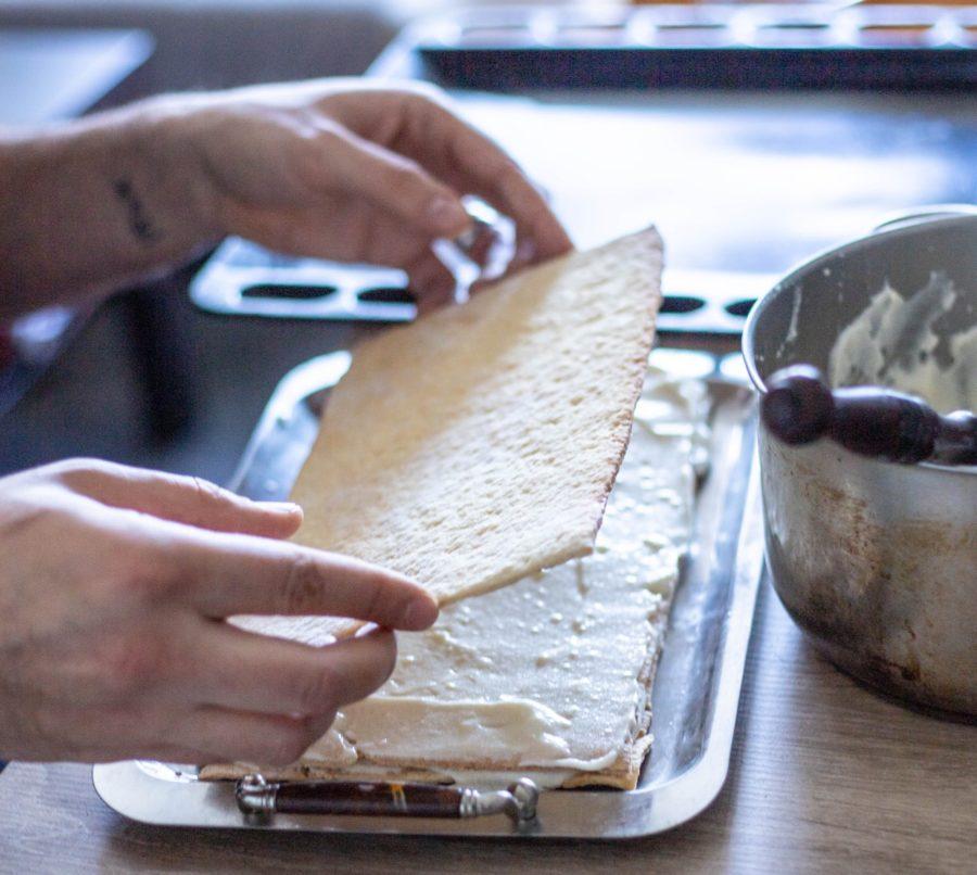 A person assembles a layered dish by placing a thin sheet of baked dough over a creamy filling on a tray. A mixing bowl and spoon are visible in the background.