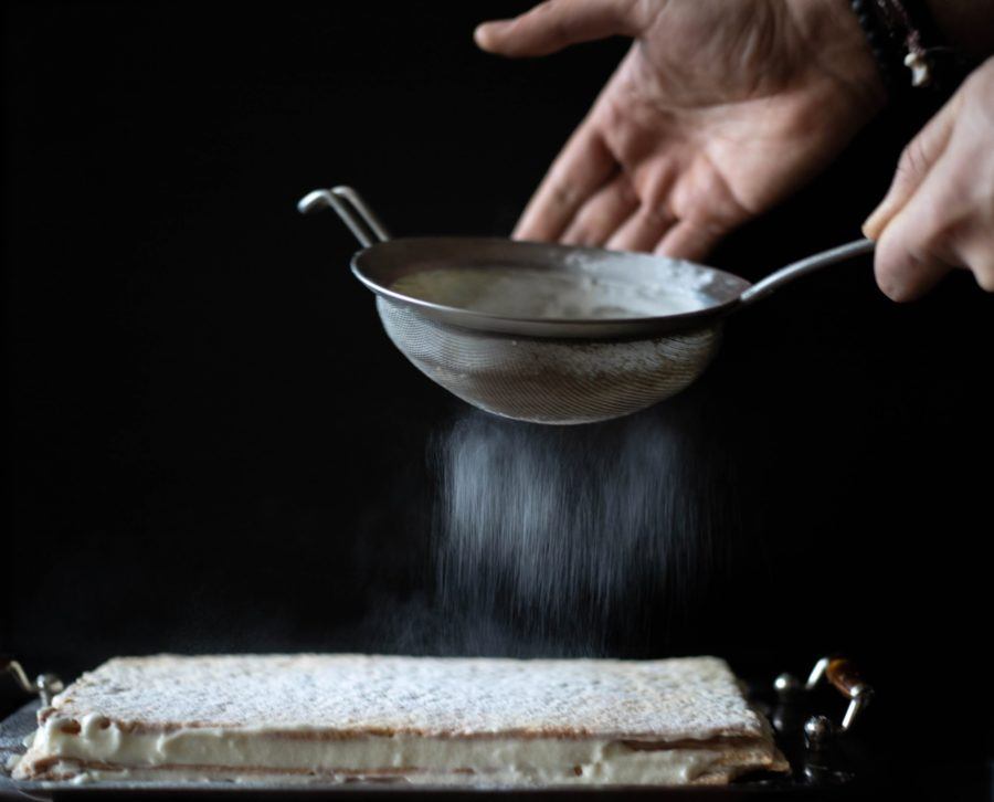 Hands are sifting powdered sugar over a layered dessert on a dark background.