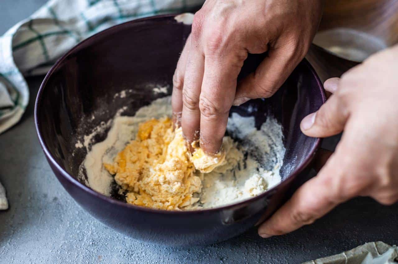 A person mixing ingredients in a dark bowl by hand, preparing dough on a countertop with a cloth in the background.