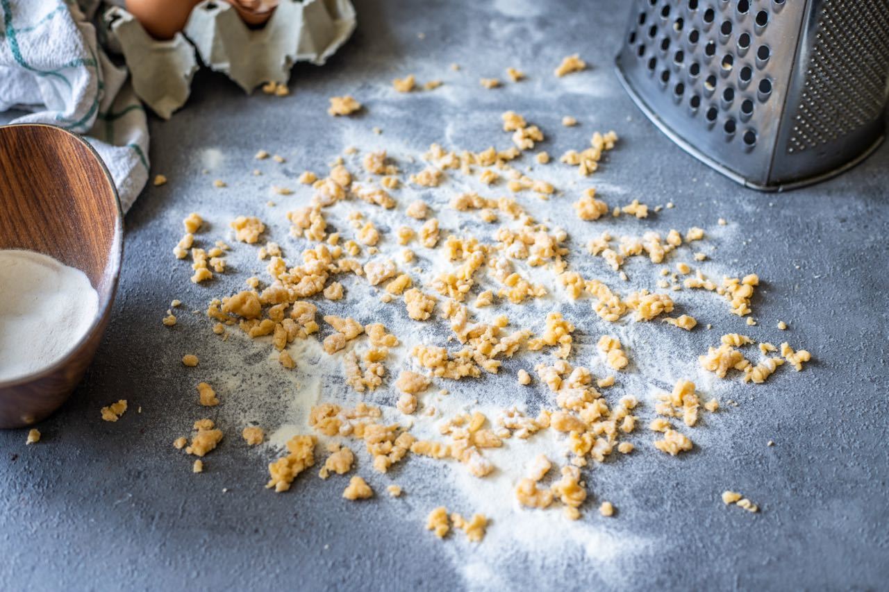Crumbled dough pieces scattered across a floured surface next to a grated utensil and a wooden bowl with white ingredients on a textured grey countertop.
