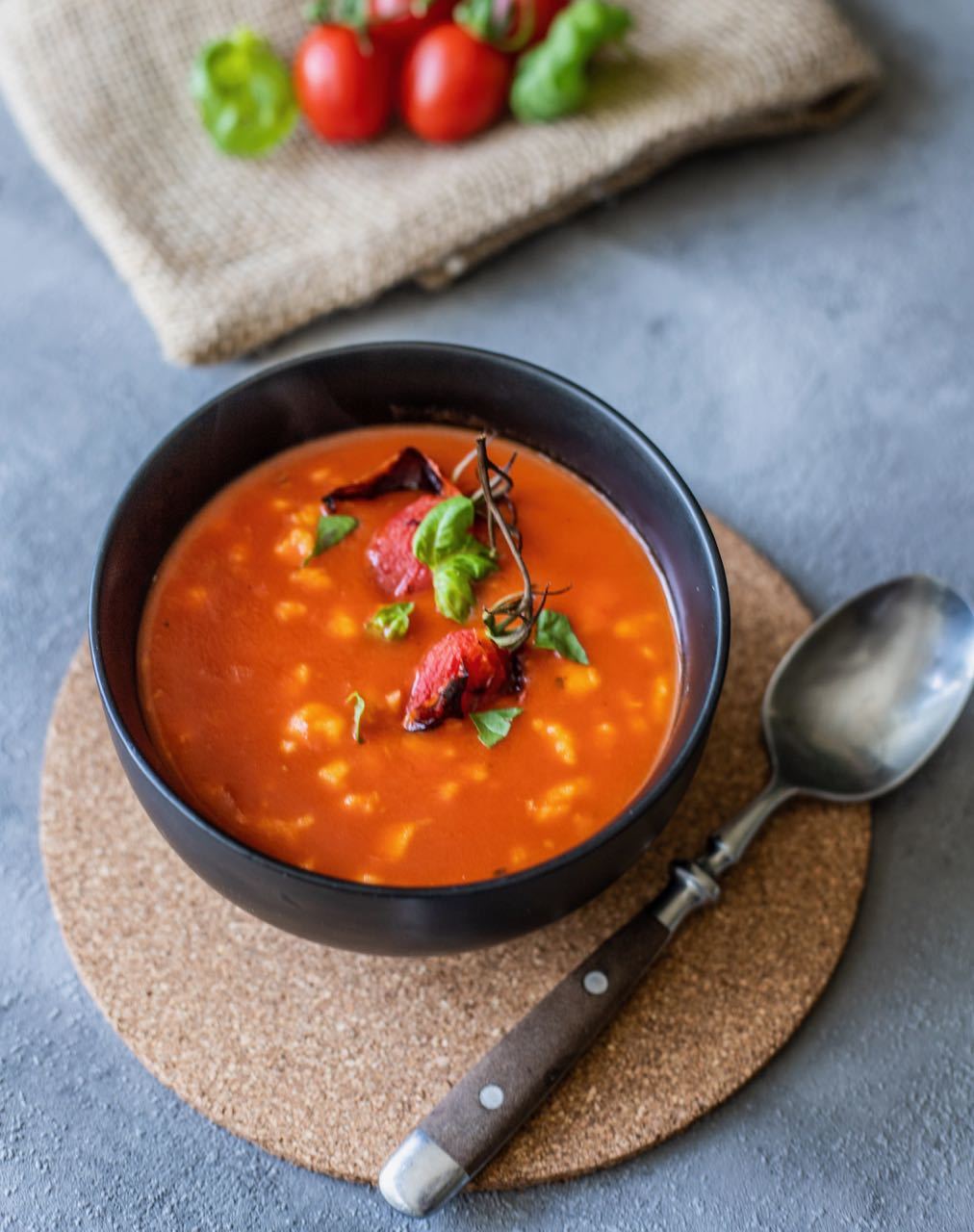 A bowl of tomato soup with basil garnish is placed on a cork mat. A spoon lies beside the bowl. Fresh tomatoes and basil leaves are in the background on a burlap cloth.