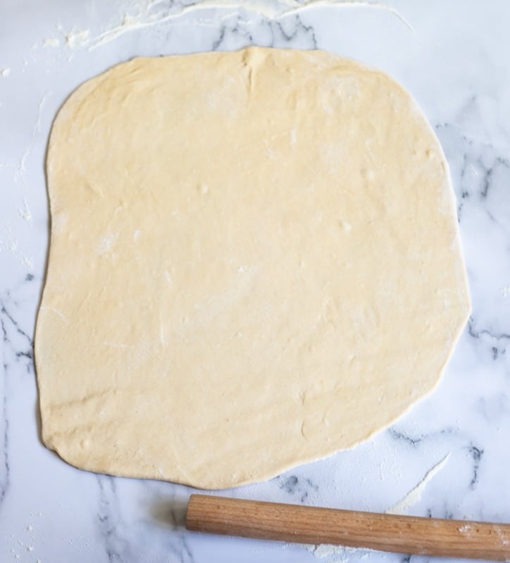 A rolled-out piece of dough on a marble surface with a wooden rolling pin next to it.
