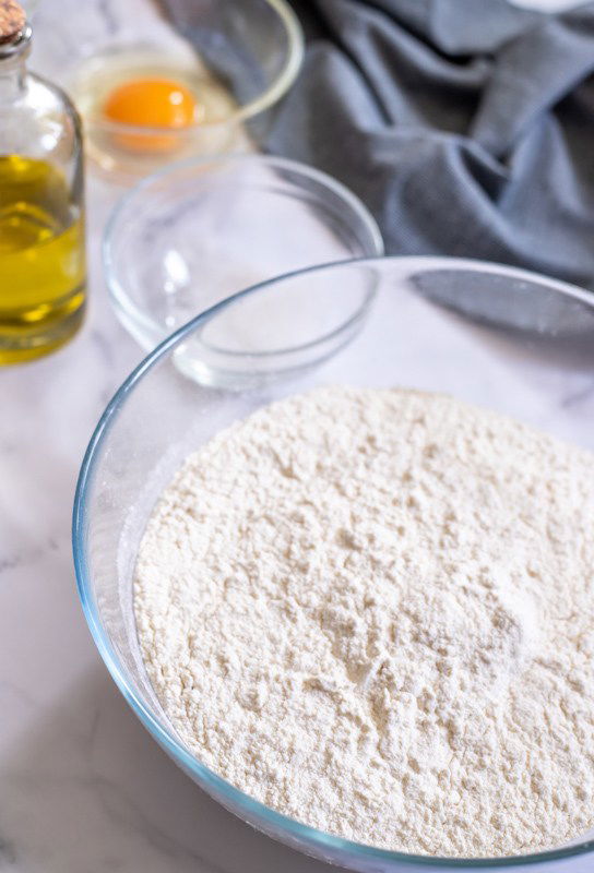 A glass bowl filled with flour, an egg in a small bowl, a bottle of olive oil, and a gray cloth are placed on a marble countertop.