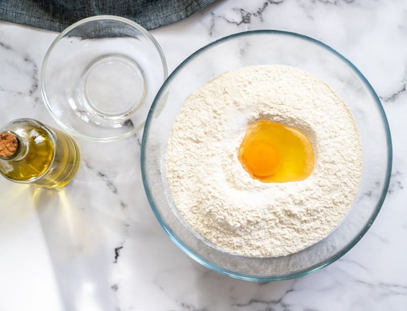 A glass bowl containing flour with a cracked egg in the center. Near the bowl are a small empty glass dish and a bottle of olive oil on a marble surface.