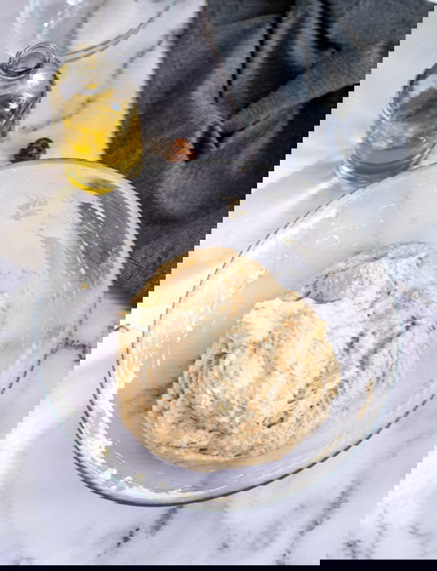 A glass bowl with dough on a marble countertop, surrounded by a bottle of oil, salt in a small container, and a gray cloth.