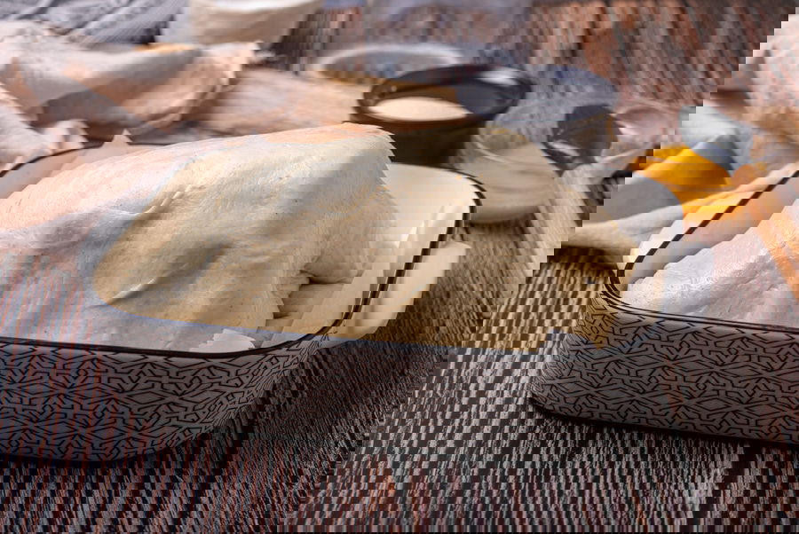 A loaf of bread dough rising in a rectangular patterned dish on a wooden surface, with various baking tools and ingredients nearby.