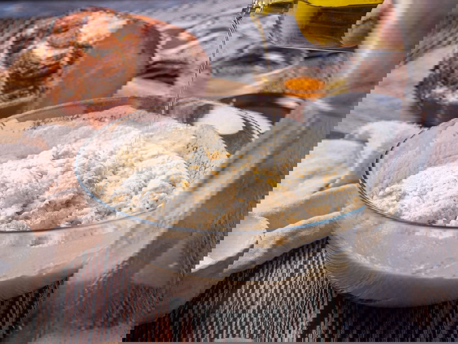 A close-up of a bowl with dough mixture as olive oil is being poured in. A piece of cooked meat and an egg yolk in a small bowl are in the background on a wooden table.
