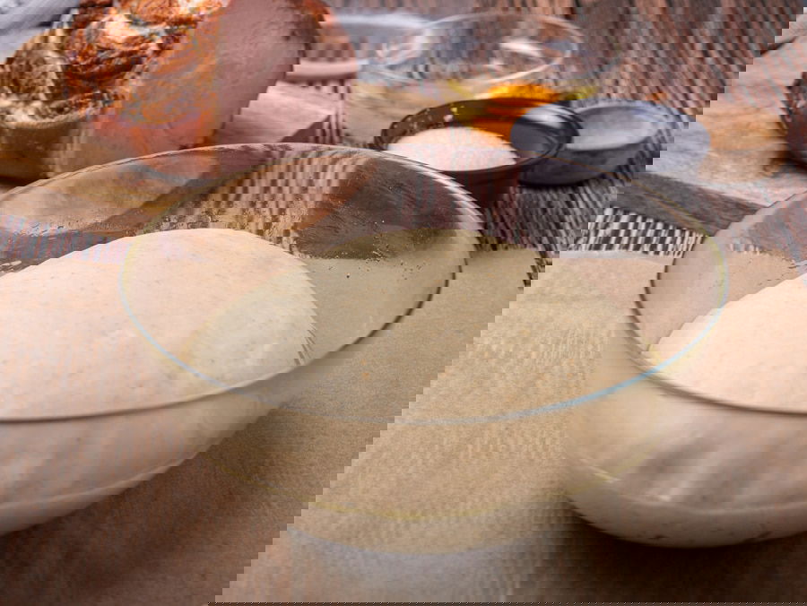 A glass bowl of risen dough on a wooden surface, with a sliced ham, a bowl of egg yolk, and a bowl of sugar in the background.