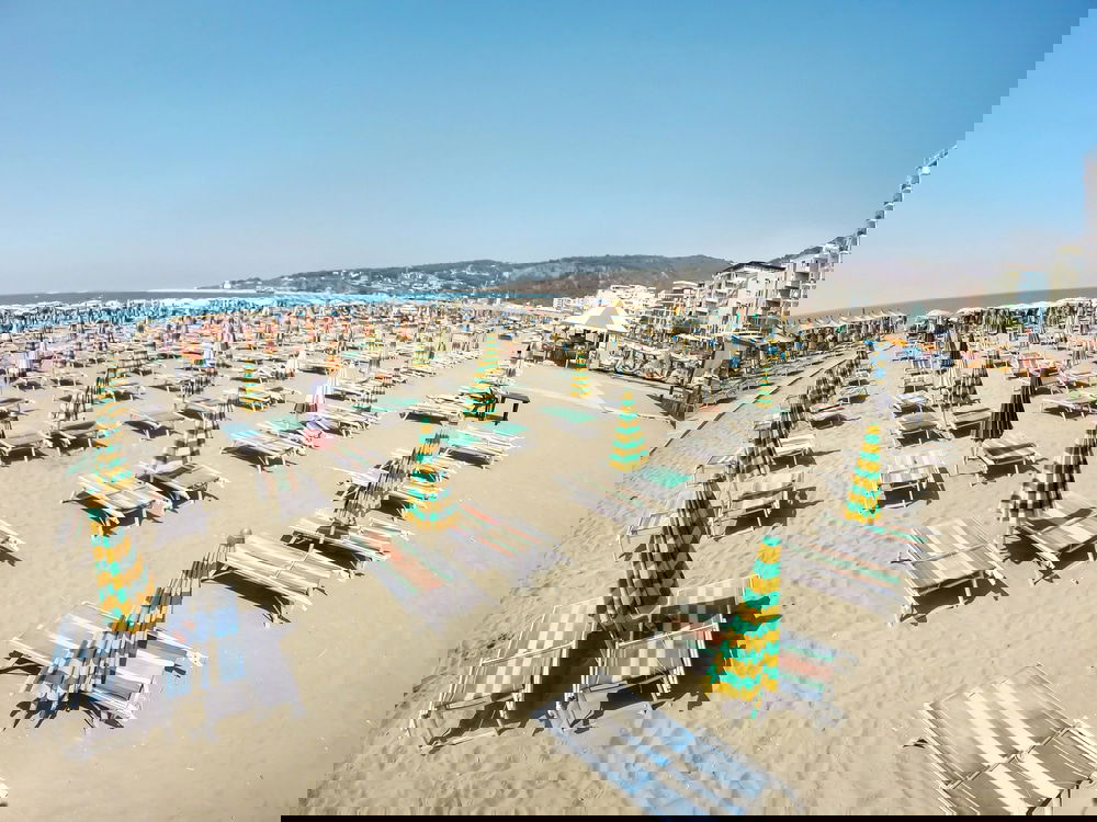 A sandy beach with rows of empty lounge chairs and closed umbrellas under a clear blue sky in Durrës beckons to visitors. The inviting scene, with buildings visible in the background, makes one wonder: Is Durrës worth visiting for its serene coastal charm?